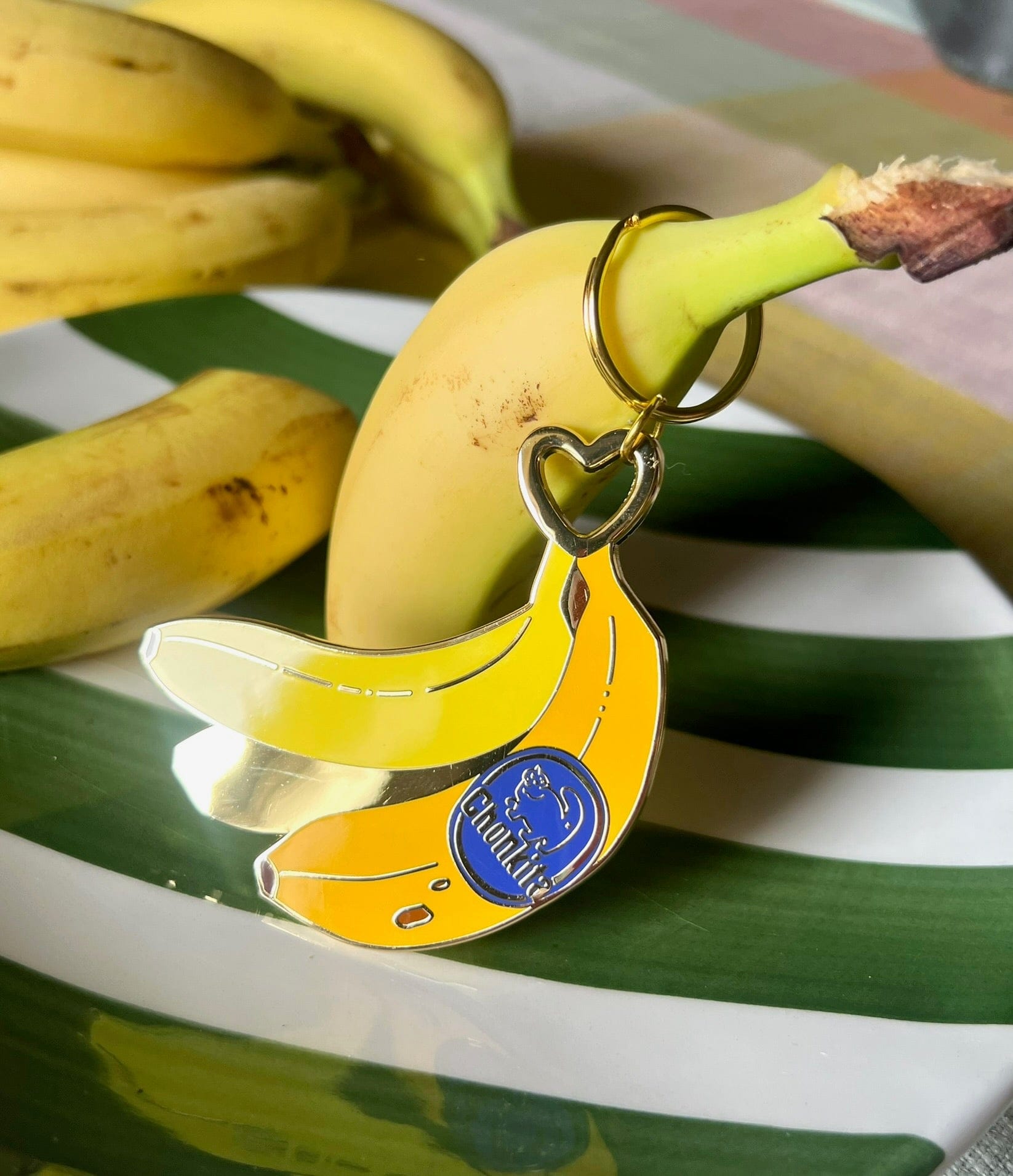 The Chonkita Banana Keyring by Hôtel Dodo, featuring a blue label, is displayed on a fresh banana with others around it on a green and white striped plate.
