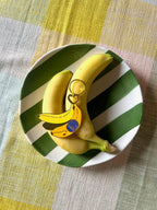 Two bananas and a Hôtel Dodo Chonkita Banana Keyring sit on a green-and-white striped plate atop a multicolored tablecloth.