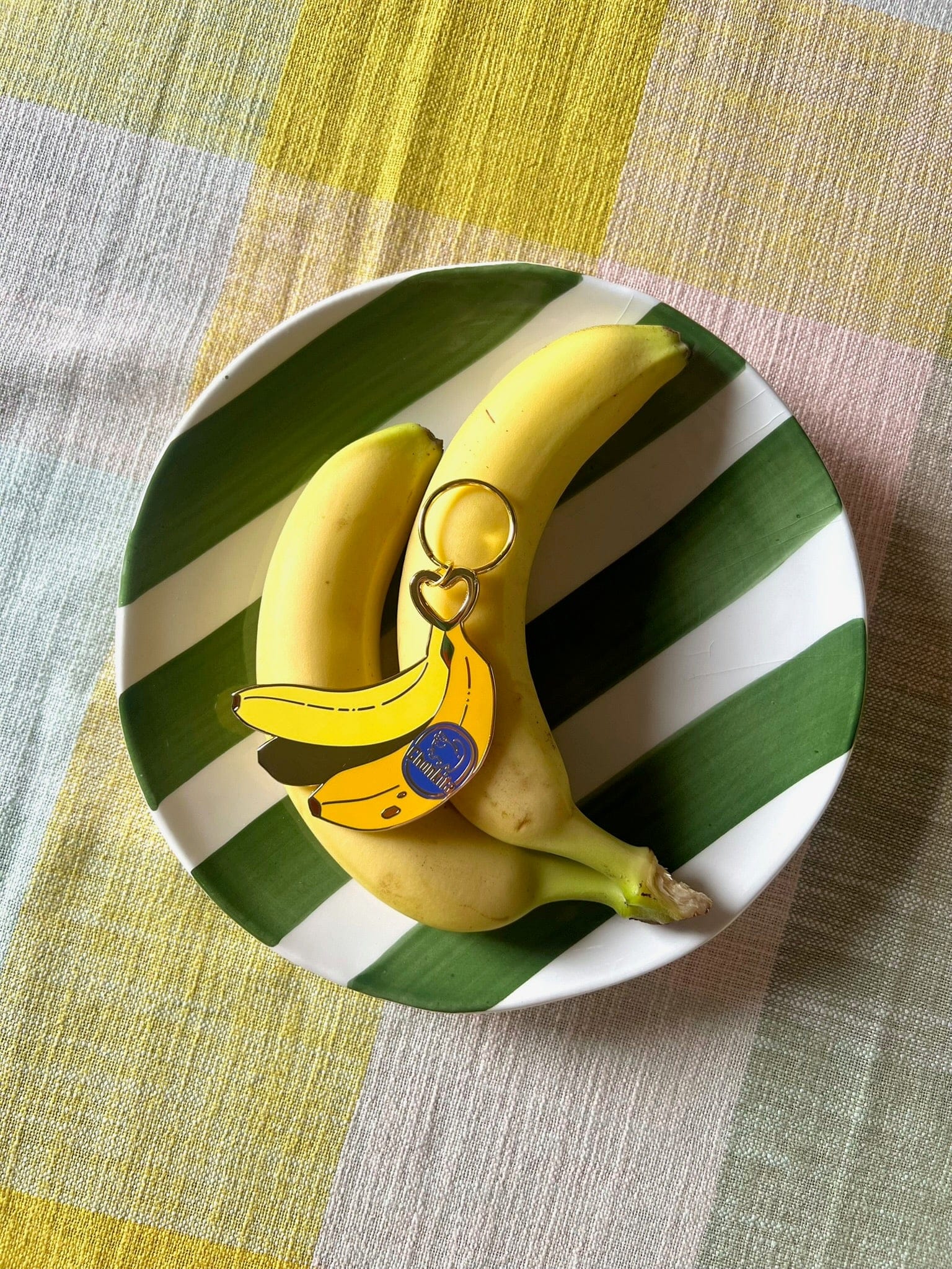Two bananas and a Hôtel Dodo Chonkita Banana Keyring sit on a green-and-white striped plate atop a multicolored tablecloth.