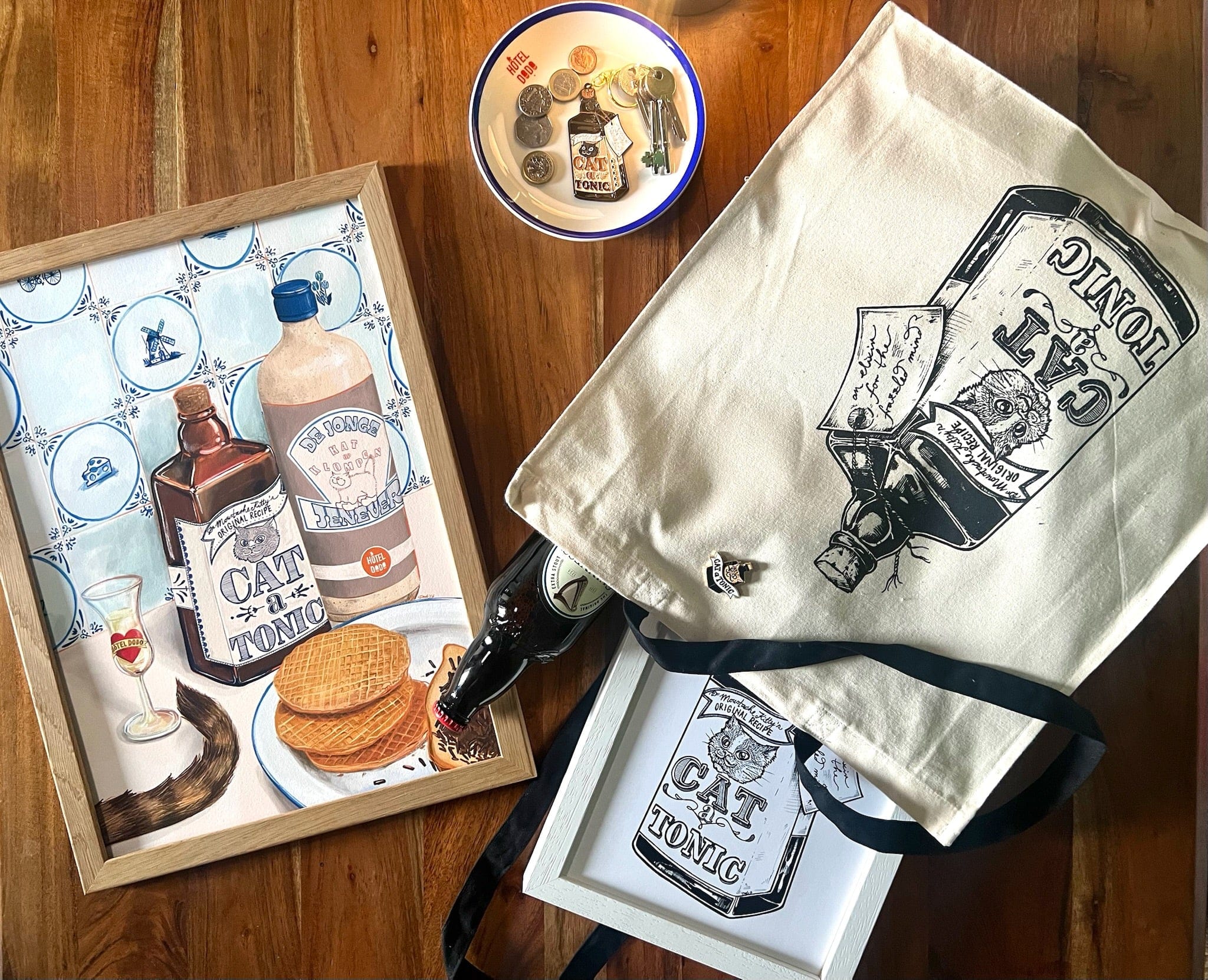 A flat lay of vintage gin-themed items, featuring the Dutch Courage Giclée Art Print by Hôtel Dodo, a tray, tote bag, bowl with coins, and a Dutch gin bottle arranged on a wooden surface.