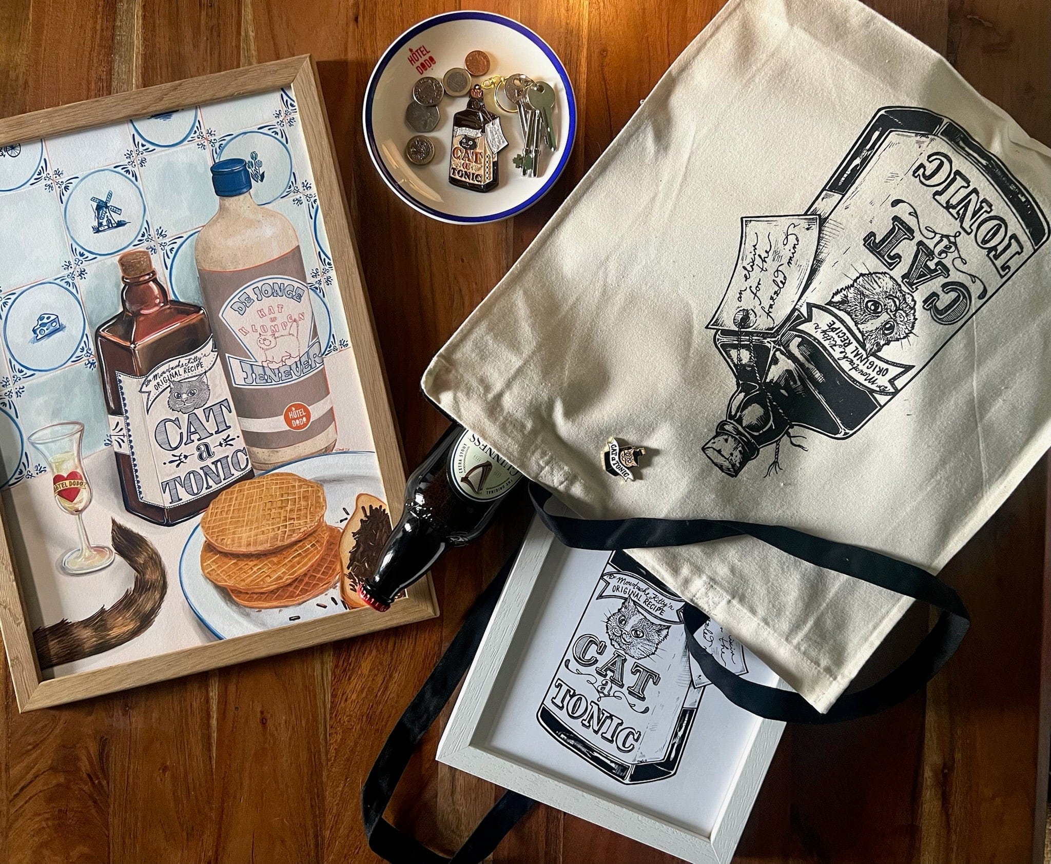 A framed painting, tote bag, and framed print by Hôtel Dodo all feature the “Dutch Courage Giclée Art Print,” inspired by Dutch gin and Jenever, showing a cat-faced bottle with coins and keys on a wooden table.