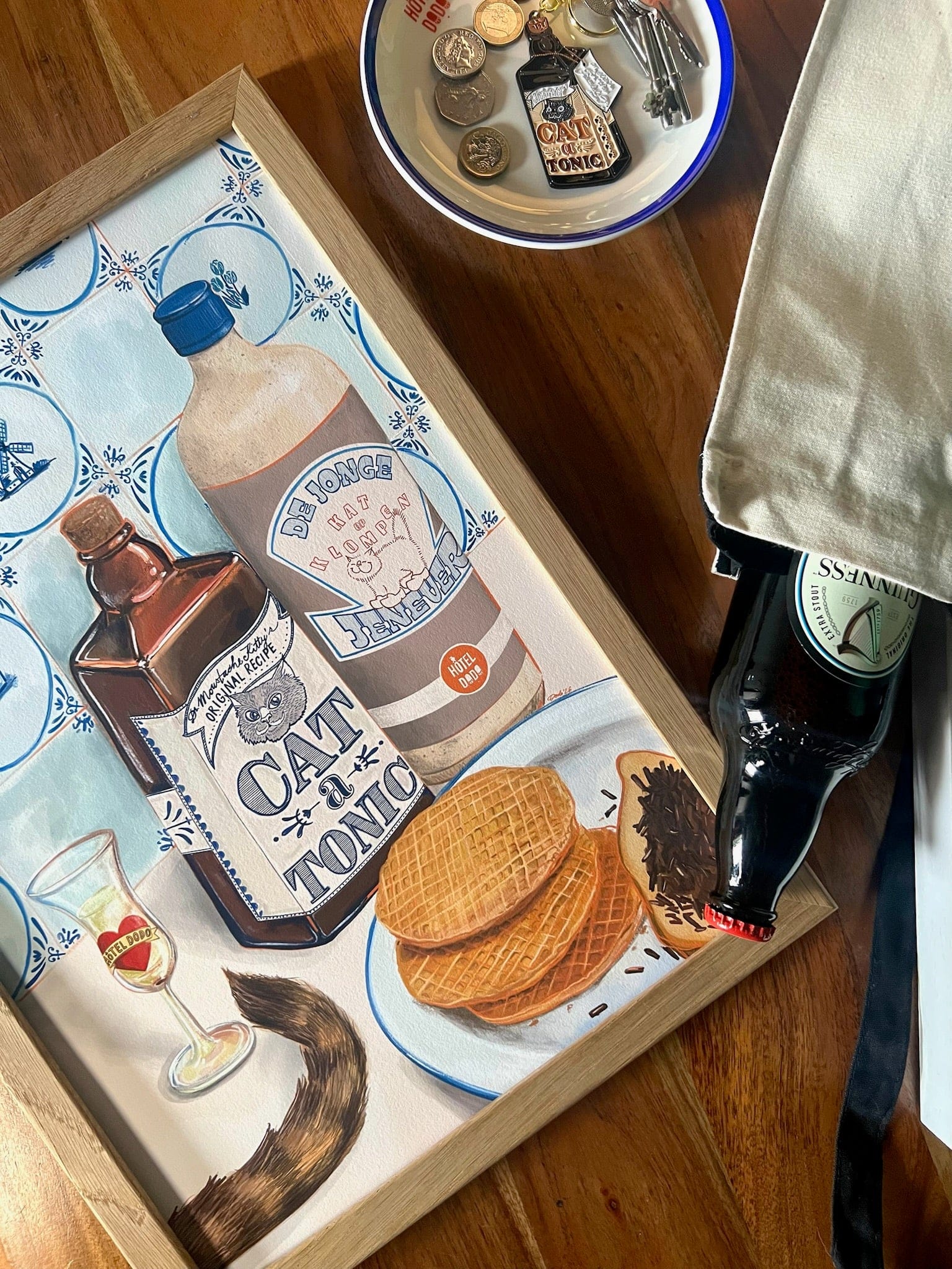 The Dutch Courage Giclée Art Print by Hôtel Dodo features stroopwafels, Cat Tonic bottles, Dutch gin, a drink, a cat’s tail, coins, and a beer bottle arranged on a table.