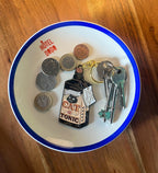A bowl labeled "Hôtel Dodo" holds several coins, a Catatonic Keyring from Hôtel Dodo, and a set of three keys on a wooden surface.