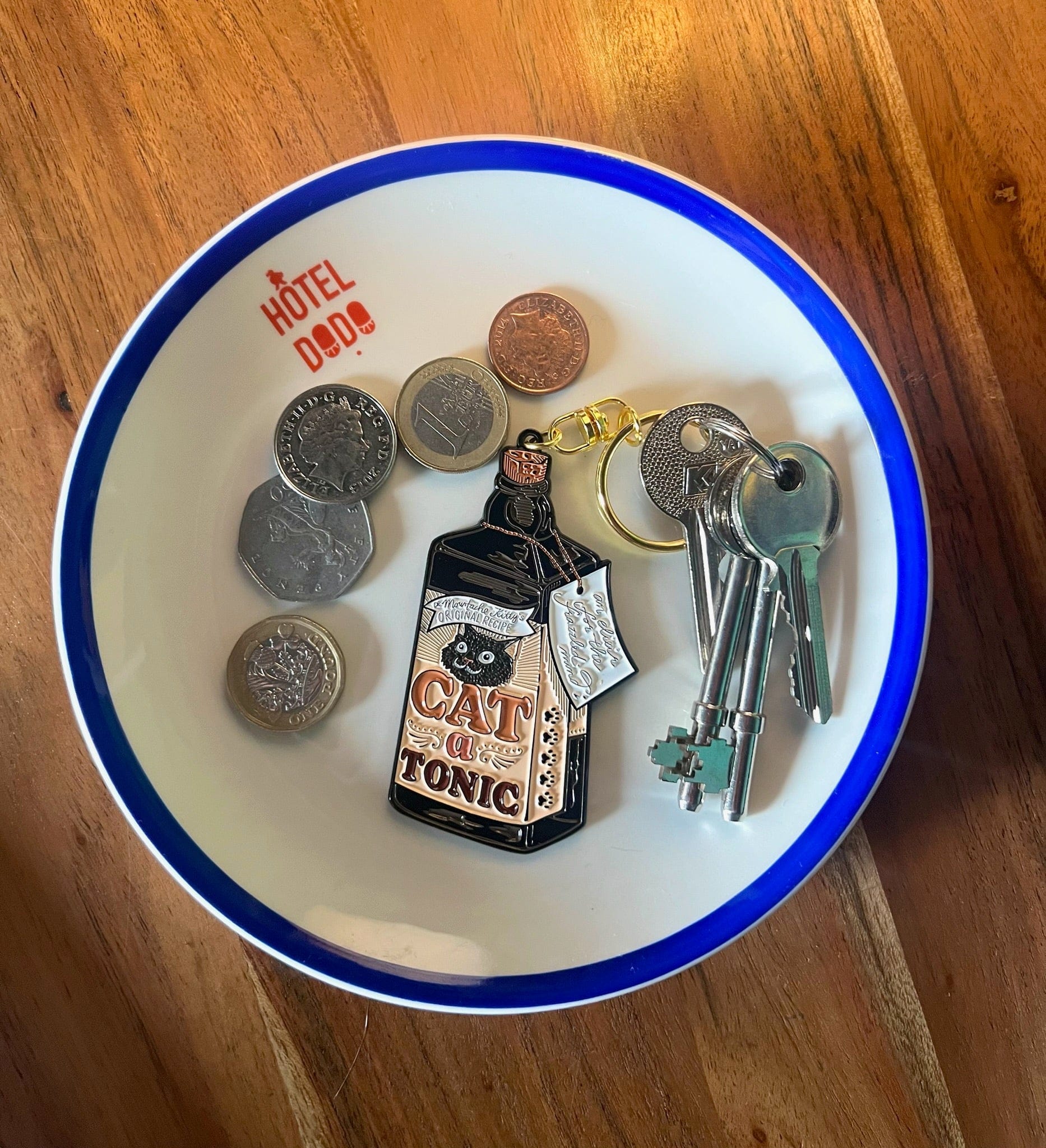A bowl labeled "Hôtel Dodo" holds several coins, a Catatonic Keyring from Hôtel Dodo, and a set of three keys on a wooden surface.