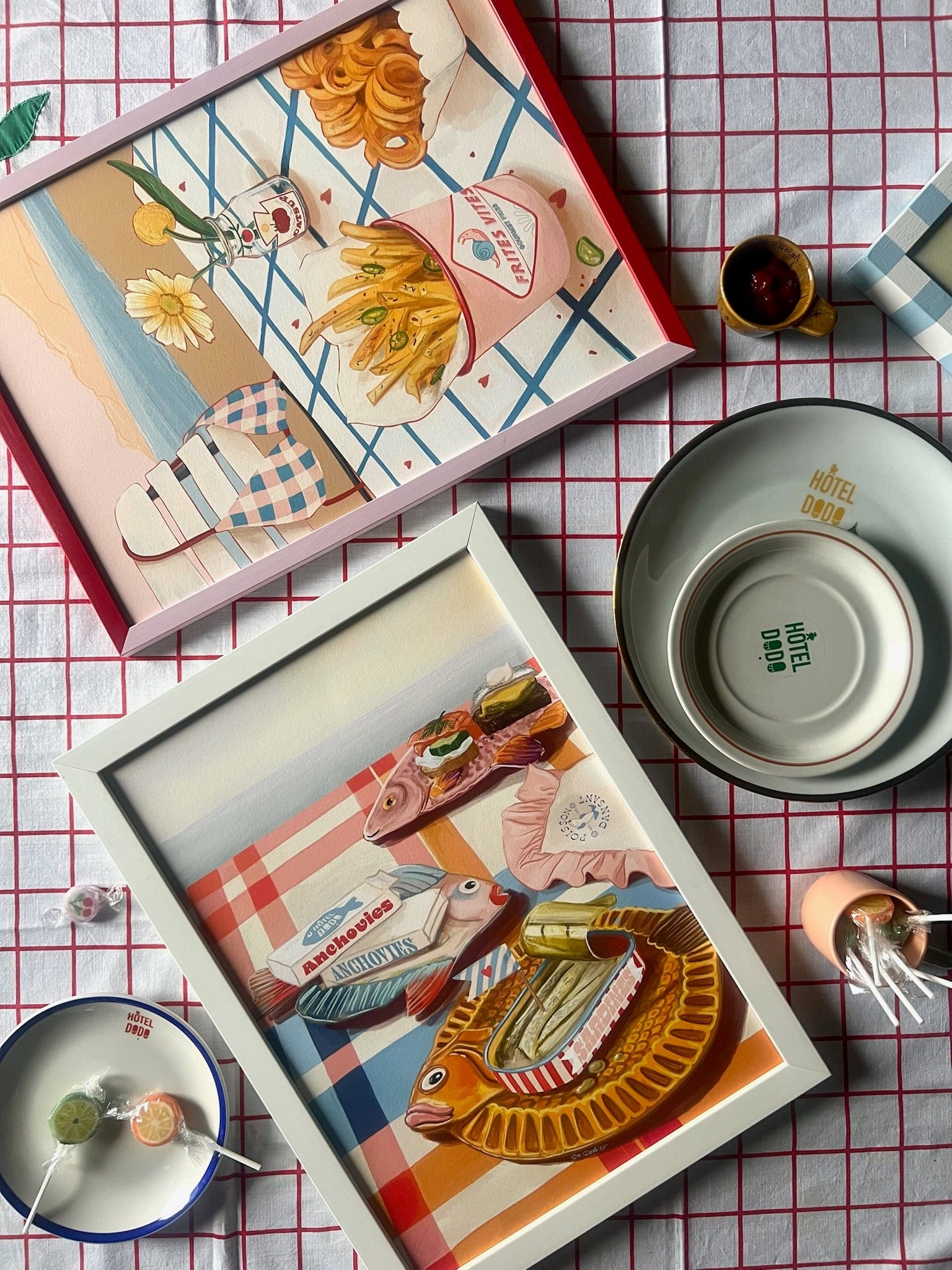 Two framed Hôtel Dodo prints—“Dishy Fishy” and a fries with ketchup piece—are arranged on a checkered tablecloth with plates and cups, creating a curated summer tablescape.