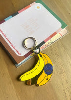 A metallic Hôtel Dodo Chonkita Banana Keyring with a blue "Goodnight" label sits on colorful cards on a wooden surface.