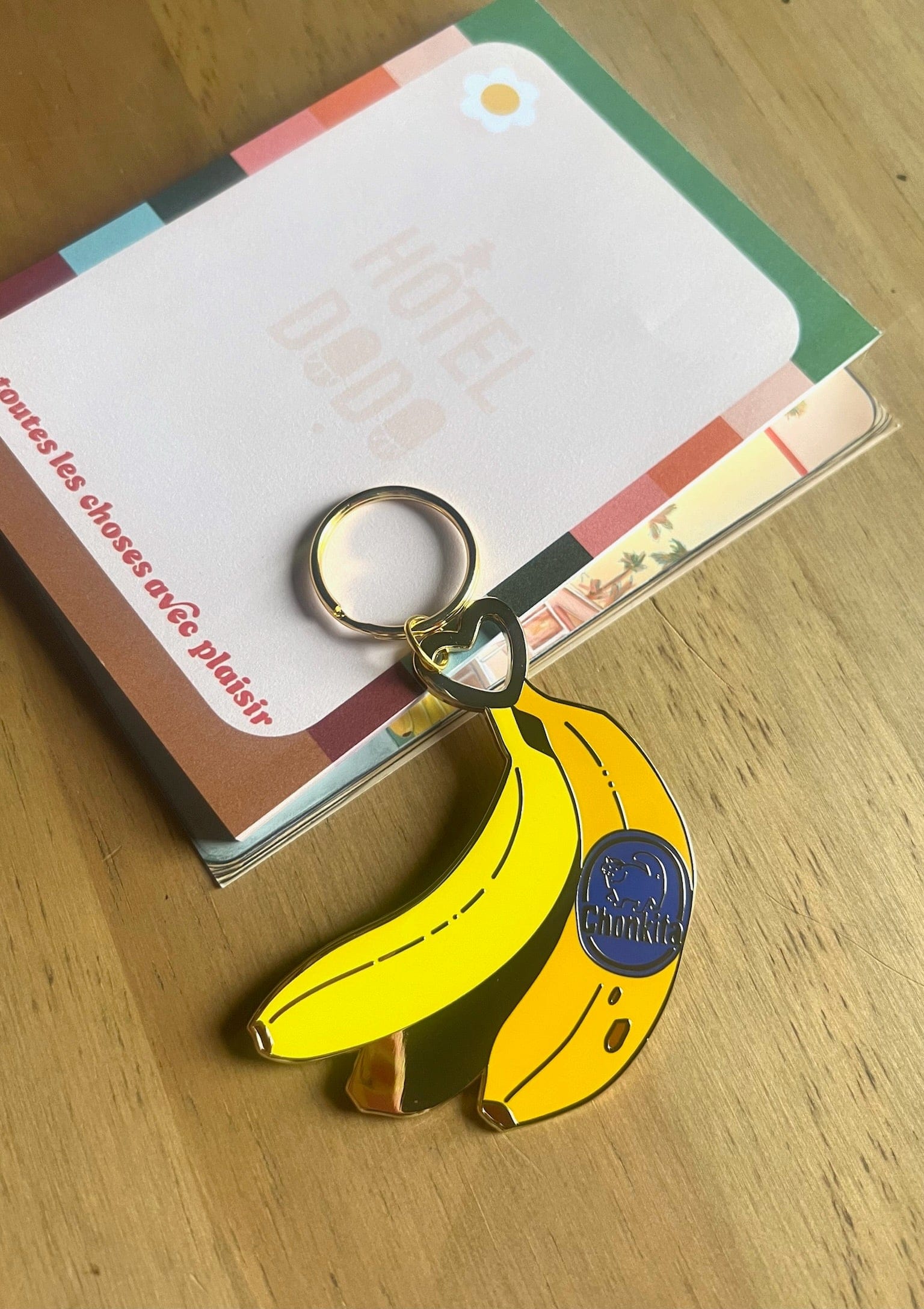 A metallic Hôtel Dodo Chonkita Banana Keyring with a blue "Goodnight" label sits on colorful cards on a wooden surface.
