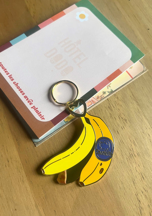 A metallic Hôtel Dodo Chonkita Banana Keyring with a blue "Goodnight" label sits on colorful cards on a wooden surface.
