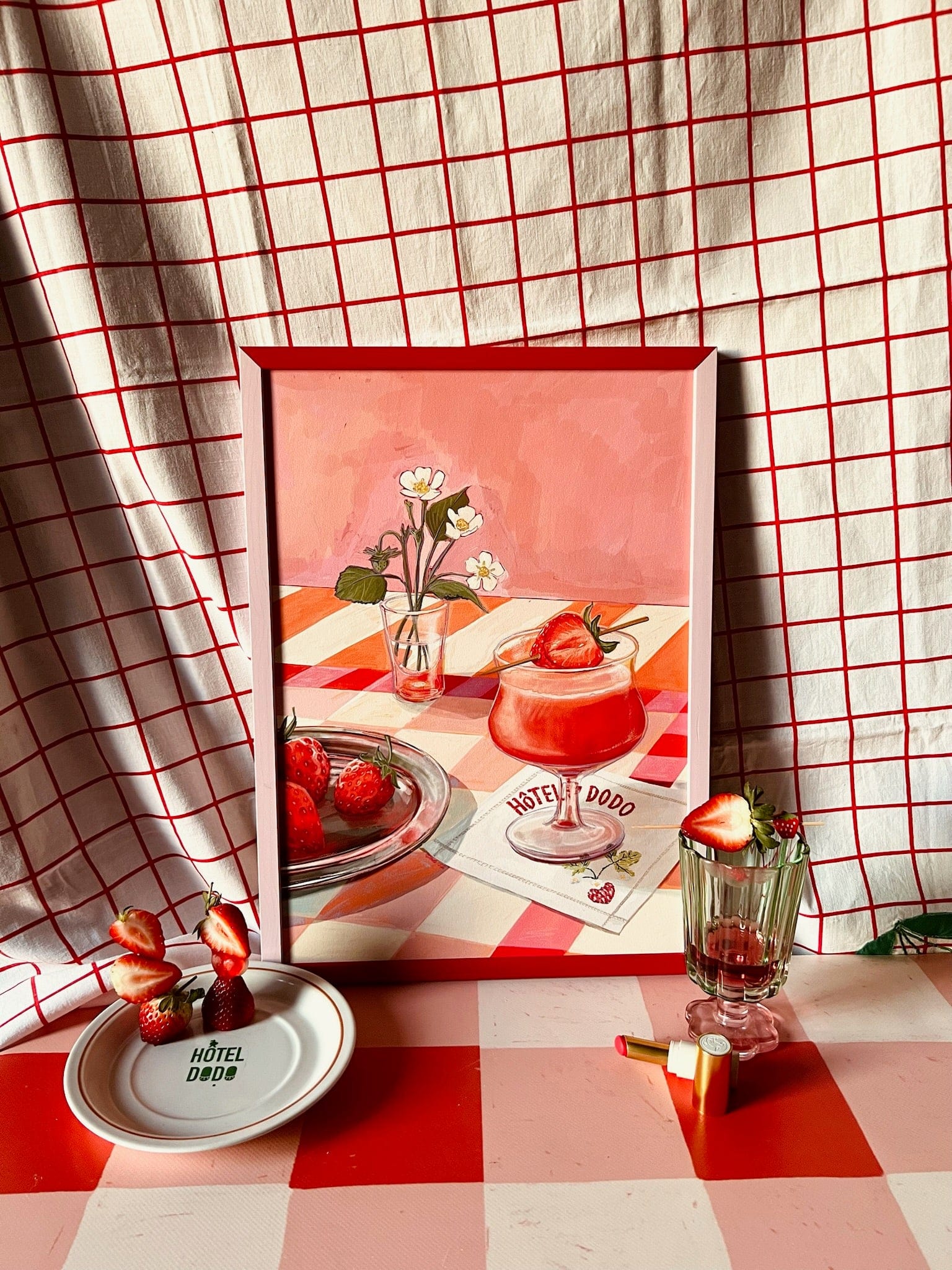 The Daiquiri Dreams Giclée Art Print by Hôtel Dodo features strawberries, flowers, and drinks on a checkered table, set against a red-and-white grid fabric with real strawberries and glass accents for a curated touch.