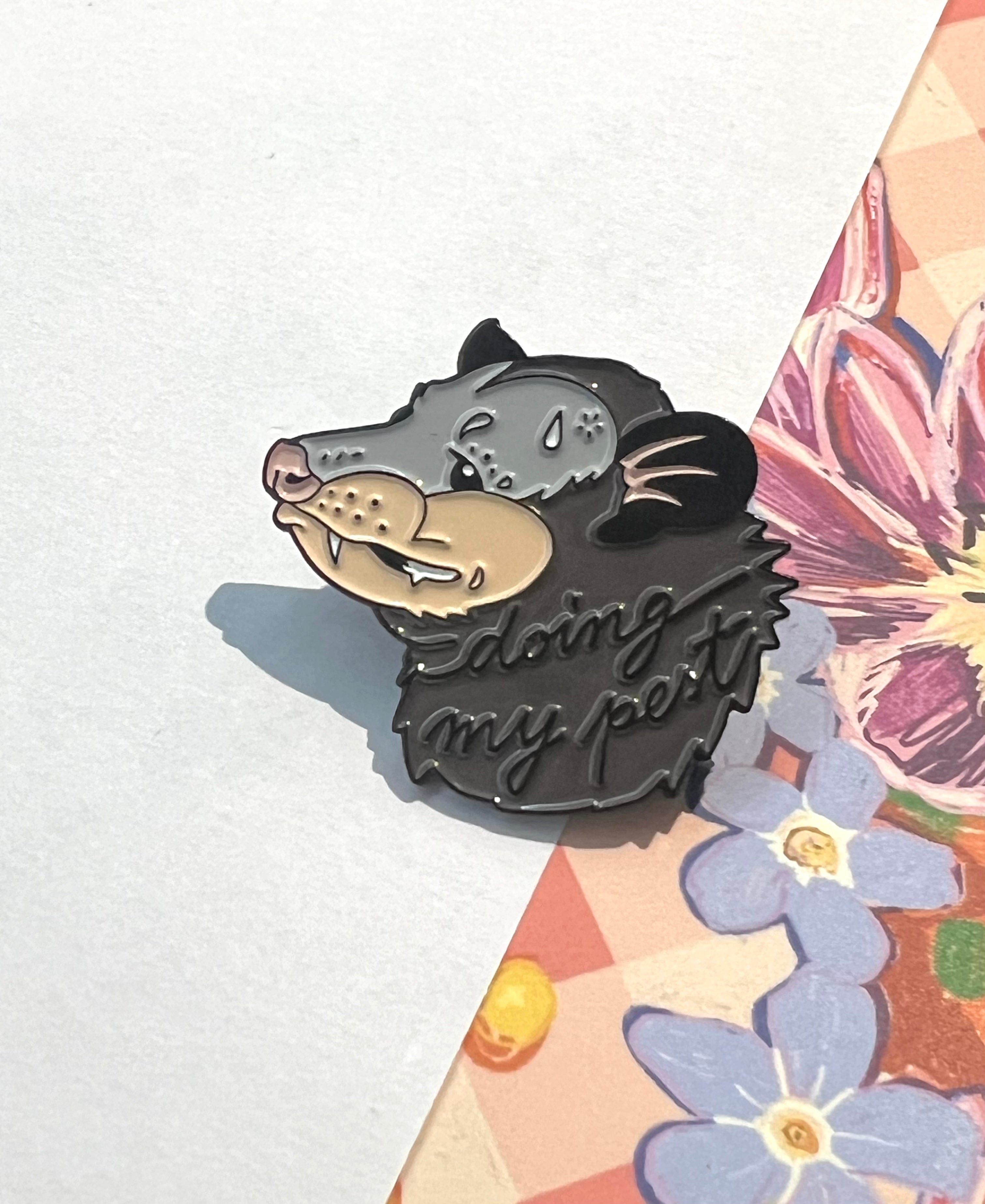 The Doing My Pest Pin by Pins by diedododa features a possum’s head with the words "doing my pest," displayed on floral-patterned paper for a touch of whimsy and luck.