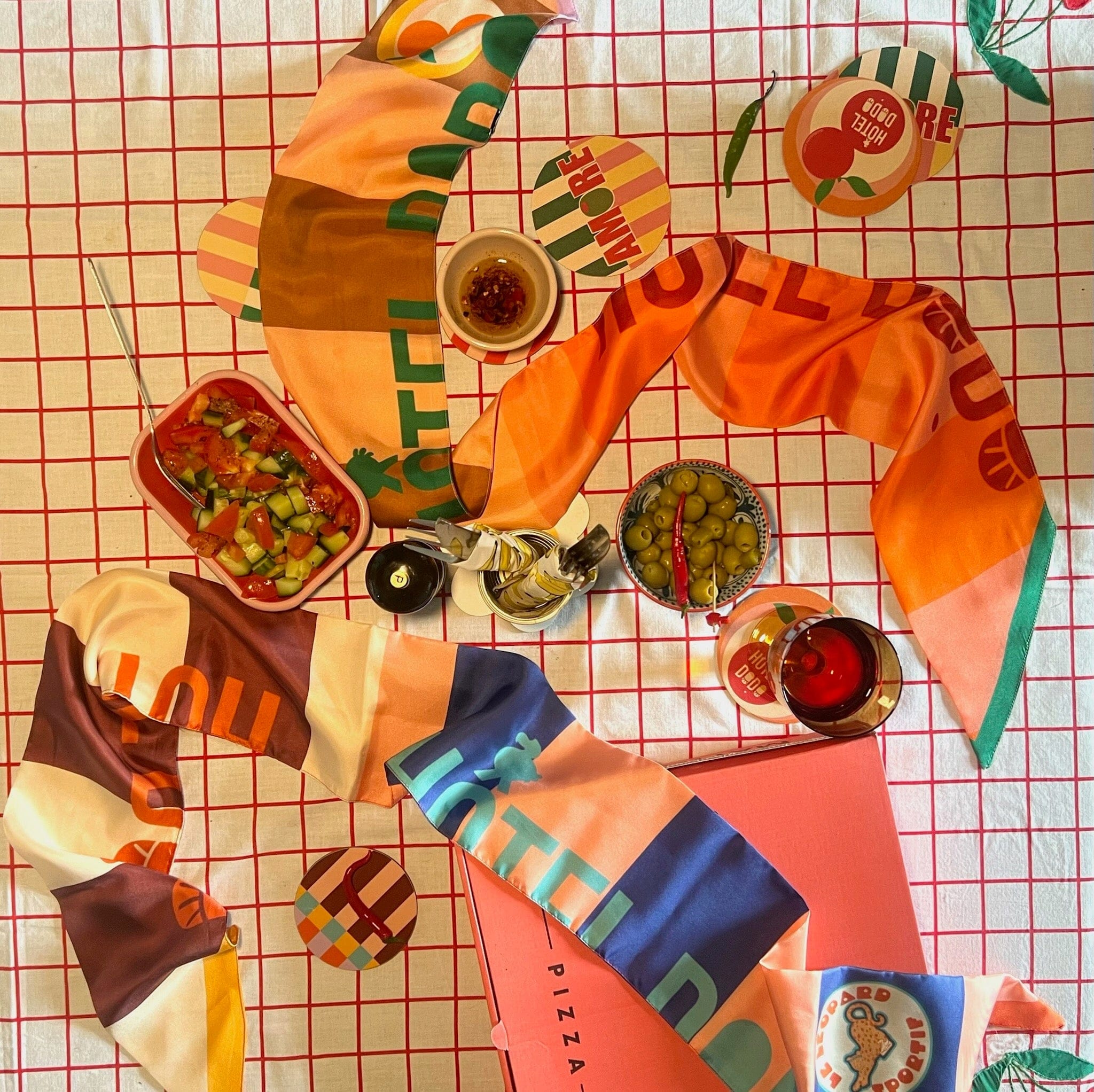 A table set with the diedododa Hotel Dodo Scarf, olives, salad, drinks, coasters, dipping sauces, and a pink pizza box on a red-and-white checkered tablecloth.