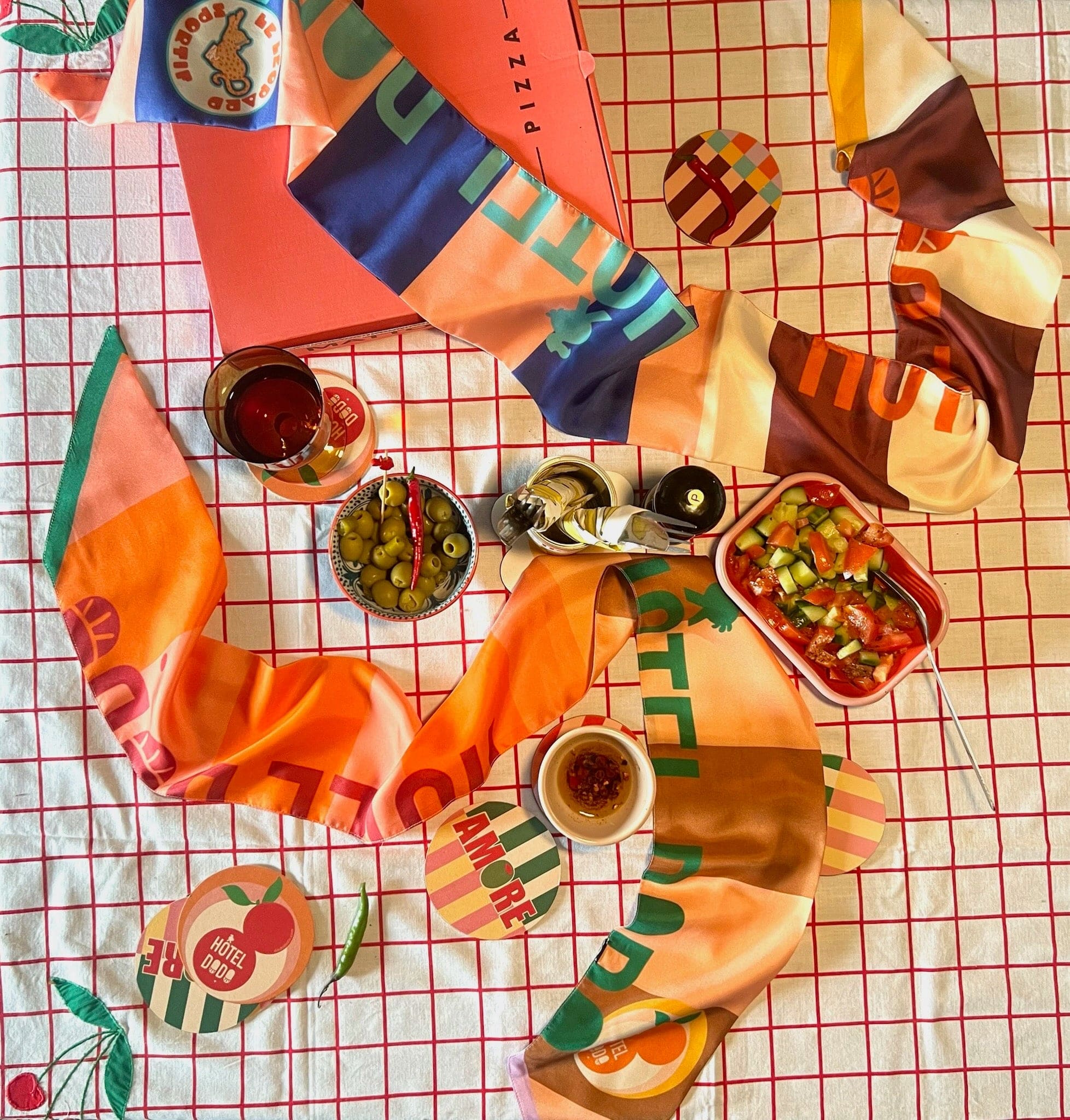Overhead view of the diedododa Hotel Dodo Scarf in silk twill, pizza box, olives, salad, sauce, and drinks arranged on a grid-patterned tablecloth.