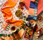 A table set with a colorful cloth, salad, green olives, a drink, chili flakes, utensils, and a pizza box features the stylish diedododa Hotel Dodo Scarf and a versatile coaster to add flair to the scene.