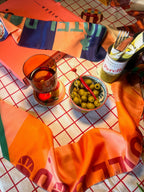 A bowl of green olives and red chili, a glass of red drink, forks in napkins, and a can on a checkered tablecloth adorned with vibrant diedododa Hotel Dodo Scarves.