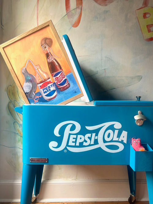 A blue Pesti Cola cooler supports a framed Kitsch Kanaveral Pesti Cola Giclée Art Print of a pelican and two soda bottles—one vintage full sugar—against a wall adorned with faint mural art.