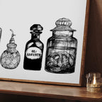 The Jellysnakes & Bellyaches Matte Art Print by Potion & Poison Bottles features a framed still life of vintage bottles, including a Victorian absinthe bottle, on wood beside a small jar with a lit candle.