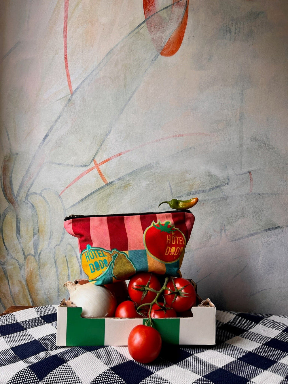 The Hôtel Dodo Fruity Pick ‘n’ Mix Pouch is placed behind a box of assorted vegetables, including tomatoes and an onion, on a checked tablecloth with a pastel-hued mural in the background.