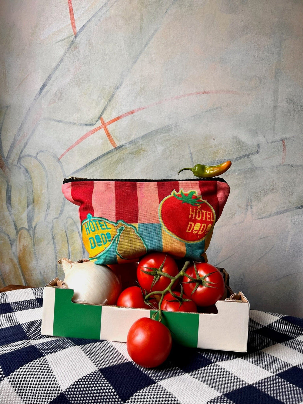 The Hôtel Dodo Fruity Pick ‘n’ Mix Pouch, striped in vibrant red and pink, is showcased on a white and green box filled with tomatoes and an onion. A chili pepper rests atop the washable fabric pouch. The backdrop includes a painted wall.