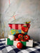 The Hôtel Dodo Fruity Pick ‘n’ Mix Pouch, striped in vibrant red and pink, is showcased on a white and green box filled with tomatoes and an onion. A chili pepper rests atop the washable fabric pouch. The backdrop includes a painted wall.