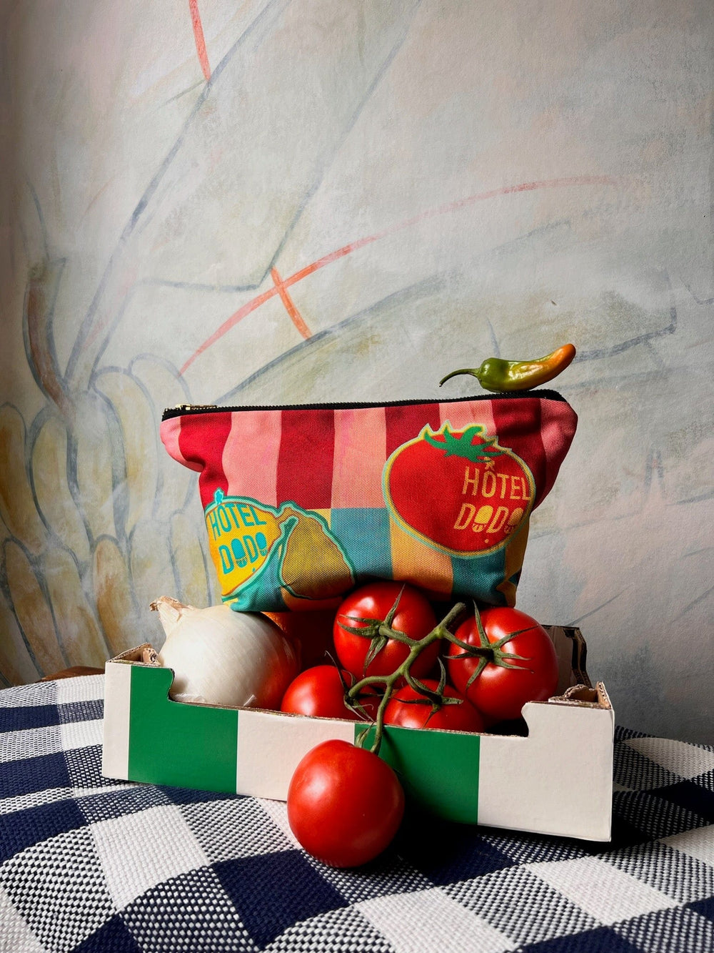 A colorful handmade Fruity Pick ‘n’ Mix Pouch by Hôtel Dodo, depicting tomatoes and peppers, is placed on a box containing fresh tomatoes and an onion, with a small pepper on top. The setup is arranged on a checkered tablecloth against a painted wall. The pouch features washable fabric for easy cleaning.