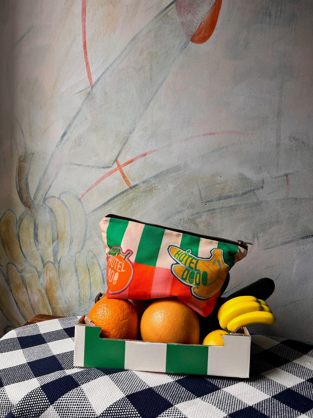 A Fruity Pick ‘n’ Mix Pouch by Hôtel Dodo sits in a box on a checkered tablecloth, with a colorful and washable fabric accessory bag placed on top, and a wall mural in the background.