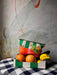 A Fruity Pick ‘n’ Mix Pouch by Hôtel Dodo sits in a box on a checkered tablecloth, with a colorful and washable fabric accessory bag placed on top, and a wall mural in the background.