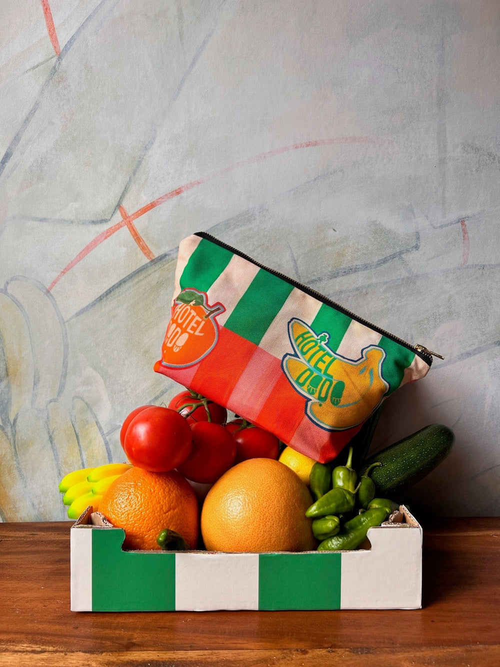 A small box filled with various fruits and vegetables, including tomatoes, oranges, bananas, and a cucumber, is placed on a wooden surface. Atop the produce sits a Hôtel Dodo Fruity Pick ‘n’ Mix Pouch made from washable fabric in a colorful, striped design.