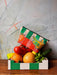 A small box filled with various fruits and vegetables, including tomatoes, oranges, bananas, and a cucumber, is placed on a wooden surface. Atop the produce sits a Hôtel Dodo Fruity Pick ‘n’ Mix Pouch made from washable fabric in a colorful, striped design.