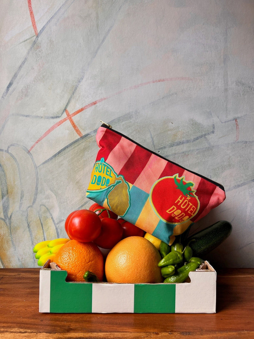 A box of various fruits and vegetables, including tomatoes, grapes, a banana, and a grapefruit, with a vibrantly printed heavyweight cotton pouch labeled "Hôtel Dodo" placed on top.