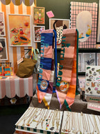 A display featuring diedododa's Hotel Dodo Scarf, along with jewelry, pins, bags, and framed art prints, all arranged on tables and walls at a market or shop booth.