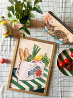 Framed artwork of a bag with groceries and a plant on a checkered tablecloth.