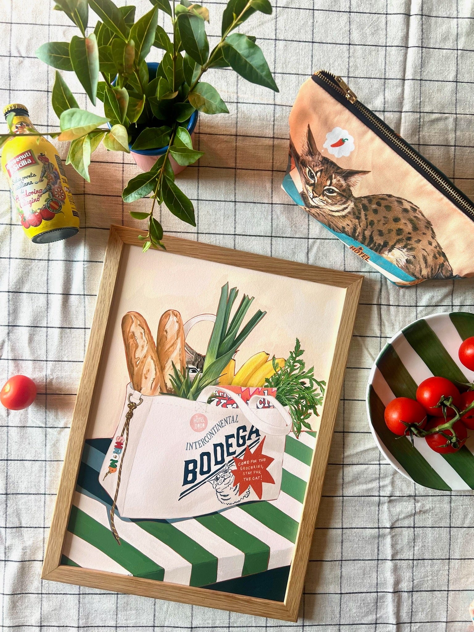Framed artwork of a bag with groceries and a plant on a checkered tablecloth.