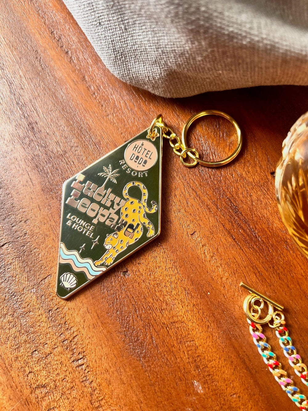 A Lucky Leopard Lounge Motel Keyring from Hôtel Dodo, featuring a golden diamond shape with a cheetah graphic and the text "Hôtel Dodo Resort, Lucky Leopard Lounge & Hotel," rests on a wooden surface beside fabric and a colorful bag charm.