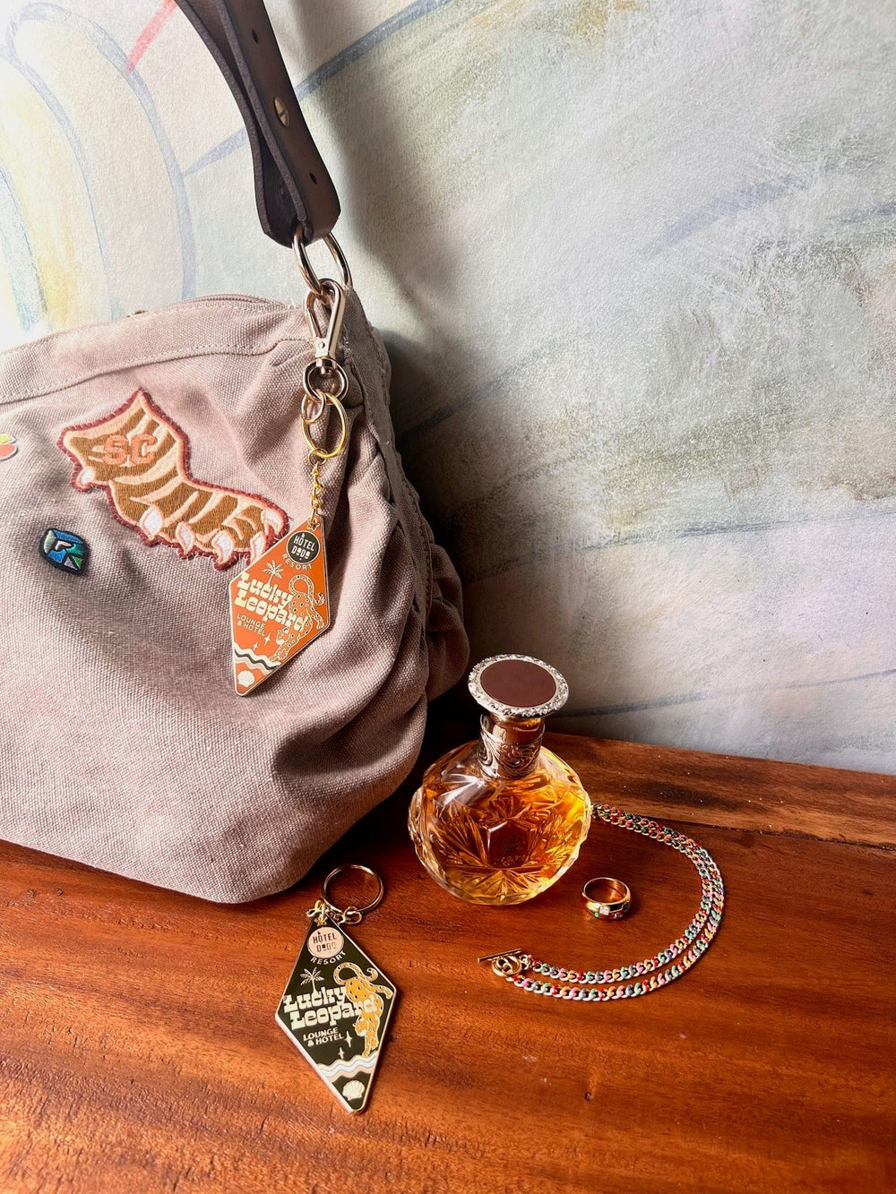 A beige bag with a Hôtel Dodo Lucky Leopard Lounge Motel Keyring, a bottle of perfume, two rings, and a silver chain are placed on a wooden surface against a light-colored wall.