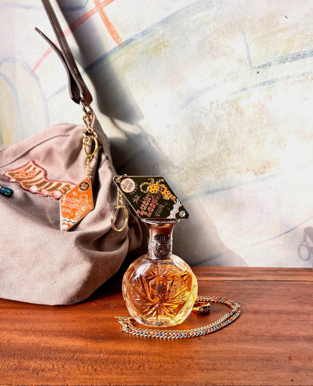 A round glass bottle of perfume with an ornate lid sits on a wooden surface next to a beige bag adorned with the Lucky Leopard Lounge Motel Keyring from Hôtel Dodo. A colorful chain and a stylish bag charm are also placed beside the perfume bottle.