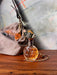 A decorative bottle of perfume with an ornate metallic stopper sits on a wooden surface. Behind it is a beige bag with a design and a Lucky Leopard Lounge Motel Keyring from Hôtel Dodo attached. The background features a painted mural.