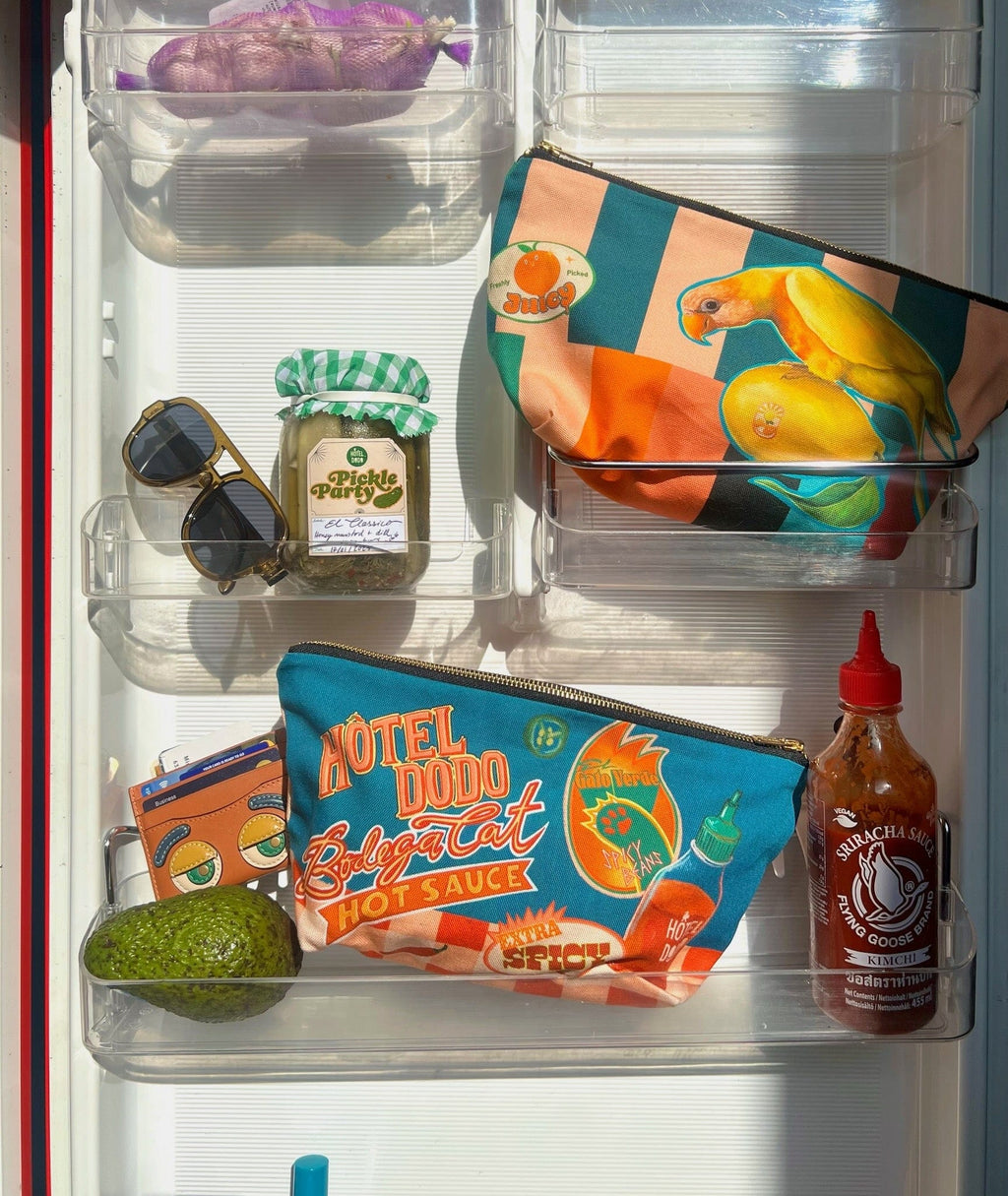 An open refrigerator door displays a Hôtel Dodo Bodega Cat Hot Sauce Pouch, a jar of pickles, sunglasses, an avocado, and a carton in the door compartments.