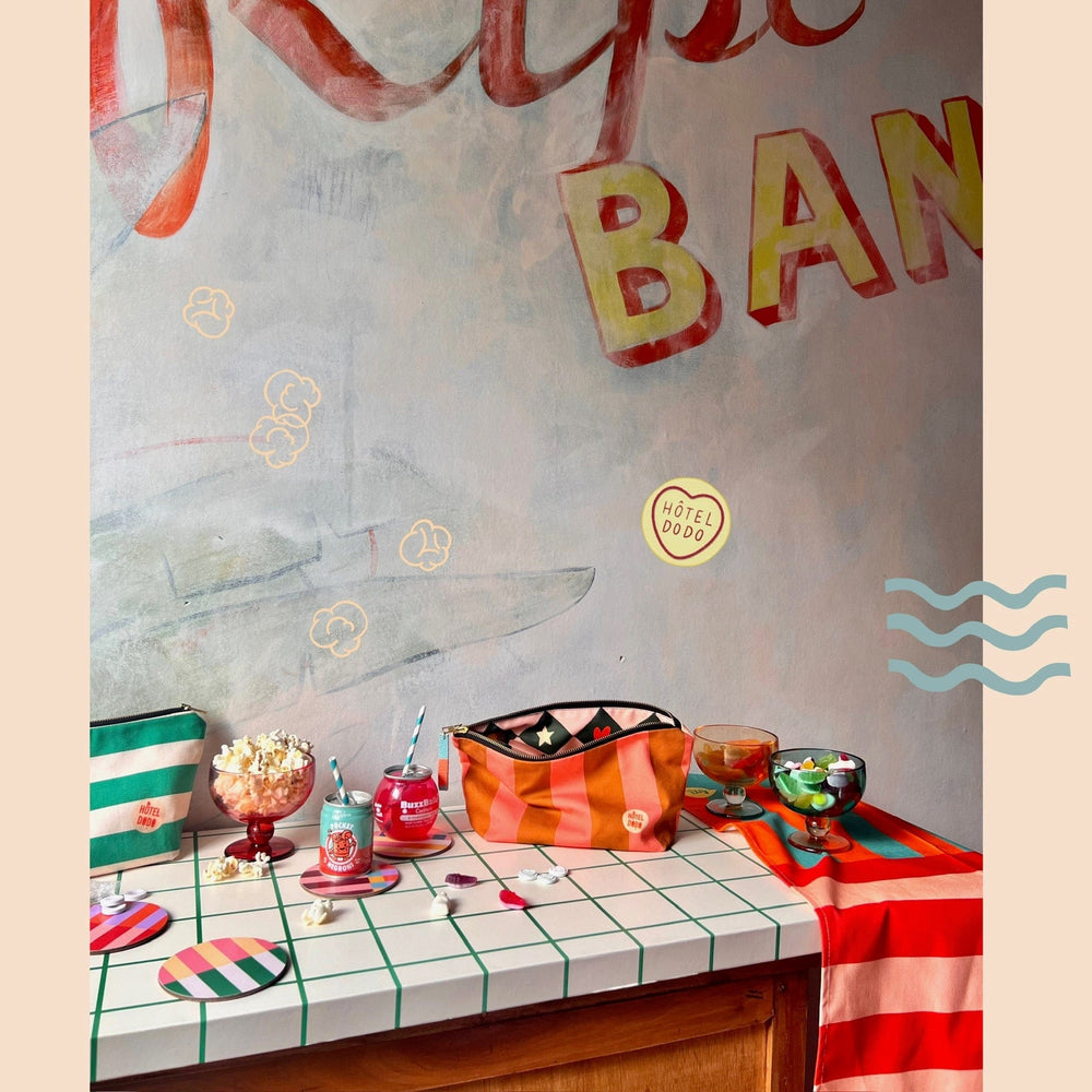 A tabletop displaying colorful items including a handmade pouch labeled "Fruity Pick 'n' Mix Pouch," striped coasters, and bowls filled with popcorn and other snacks. A mural with partial text "BAN" and the brand name "Hôtel Dodo" is visible in the background.