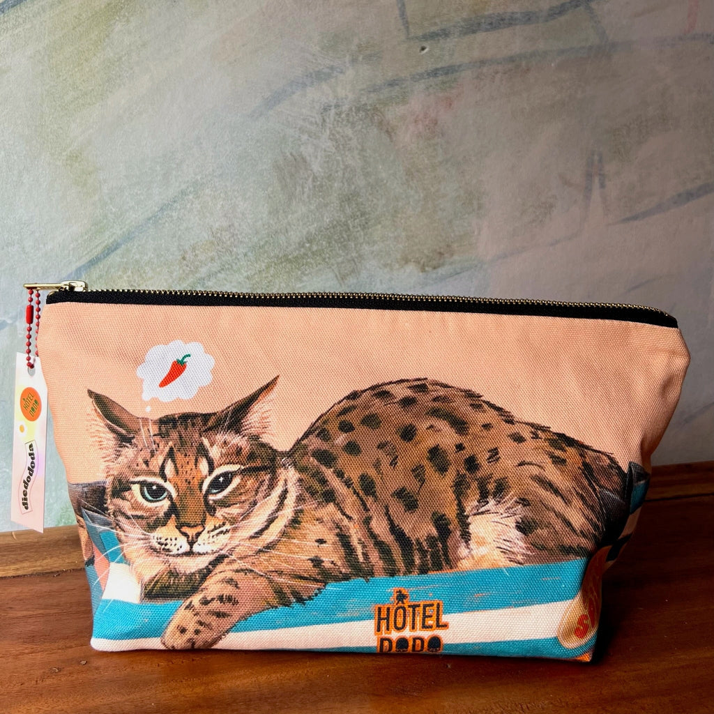 The Bodega Cat Hot Sauce Pouch by Hôtel Dodo is a rectangular cotton organiser with a lounging spotted cat and carrot thought bubble—an ideal handmade pouch for UK shoppers, shown here on a wooden surface.