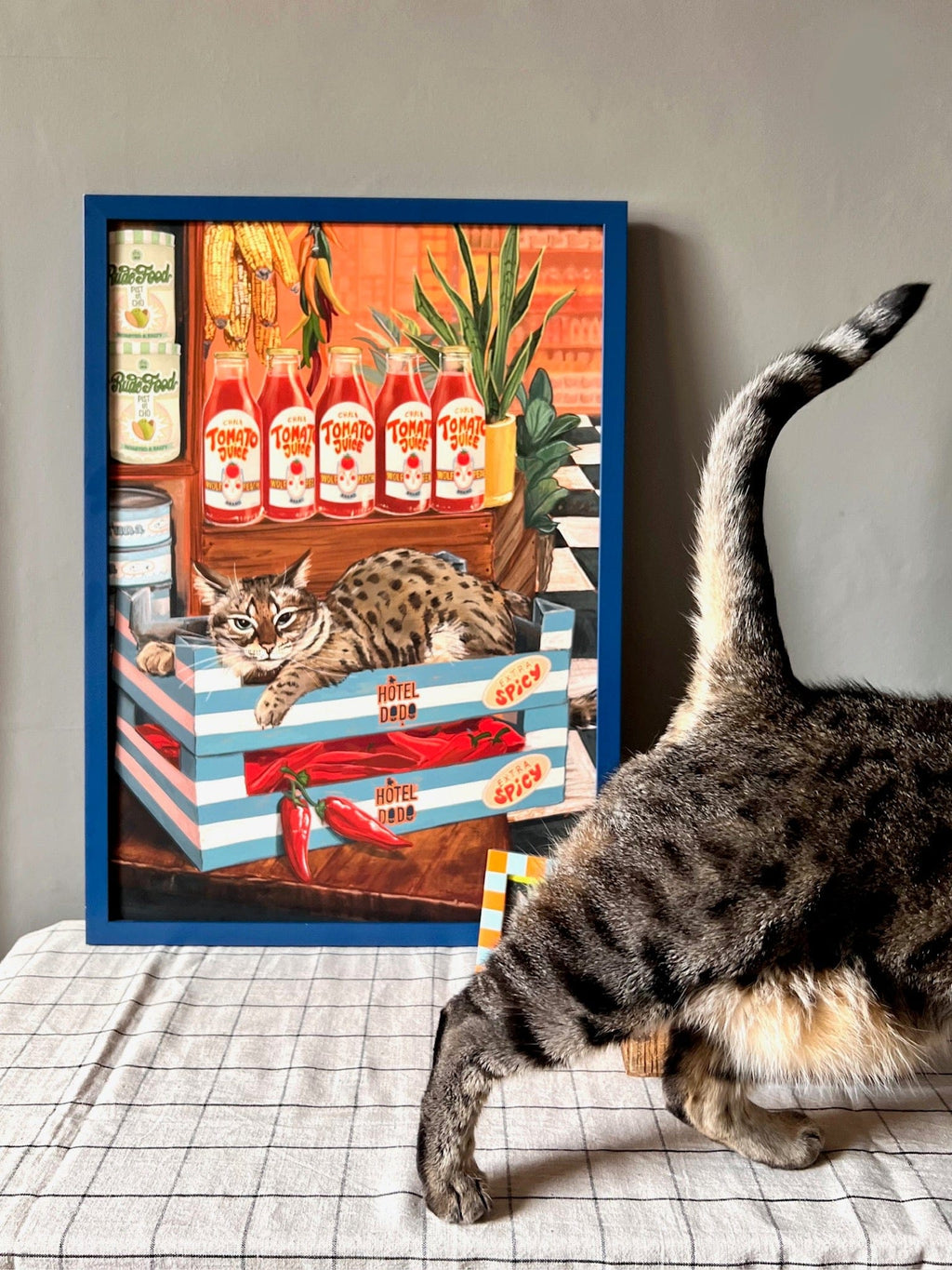 A Bodega Cat Giclée Art Print by Hôtel Dodo stands on a table with food items at the gift shop, as the hind legs of a real bodega cat pass by in the foreground.