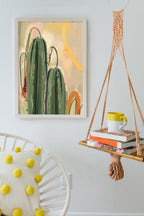 Heat Flares’ Saguaro Sundown Giclée Art Print hangs on a white wall, paired with a macramé shelf holding books and a mug. A cozy white chair accented by a yellow polka dot pillow adds comfort to the setting.