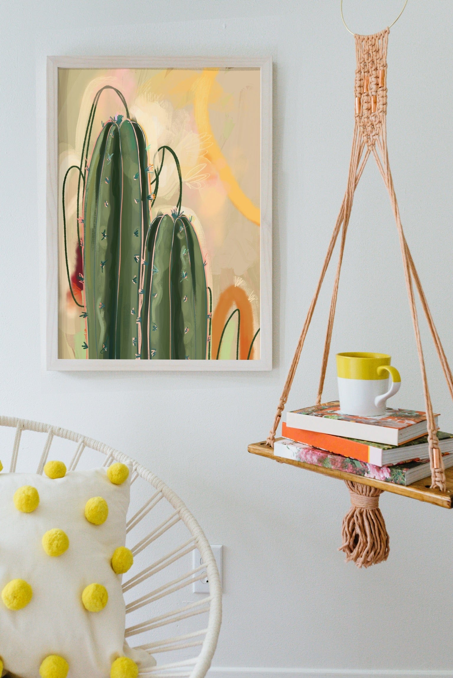 Heat Flares’ Saguaro Sundown Giclée Art Print hangs on a white wall, paired with a macramé shelf holding books and a mug. A cozy white chair accented by a yellow polka dot pillow adds comfort to the setting.