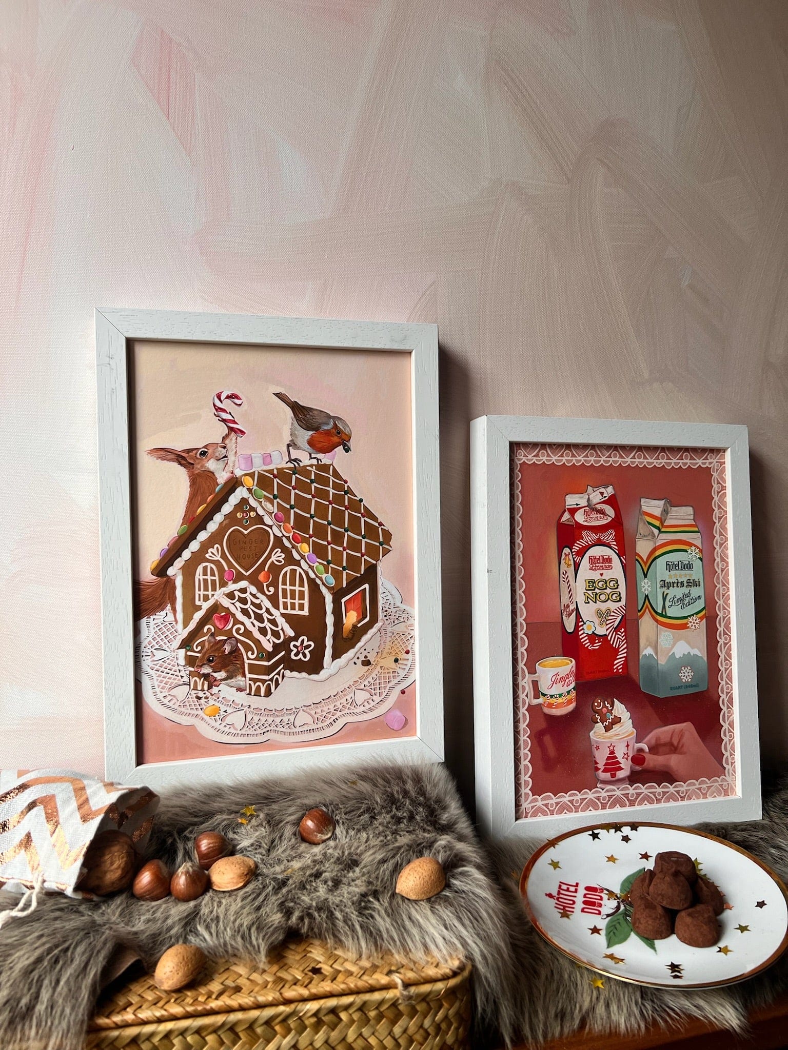 A Gingerpest House Giclée Art Print by Kitsch Kanaveral and a vintage milk carton print rest on a fur-lined basket, with nuts, chocolates, and a festive plate arranged around them for a charming holiday display.