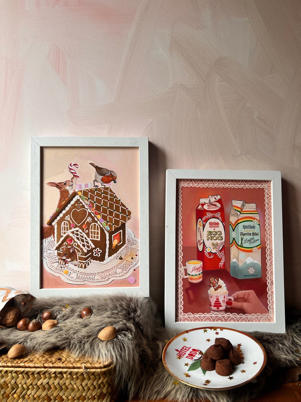 Two framed Kitsch Kanaveral x Hôtel Dodo Noggin’ The Nog Giclée Art Prints are displayed on fur with assorted nuts, chocolate truffles, and an egg nog carton from Hôtel Dodo’s festive gift shop in front.