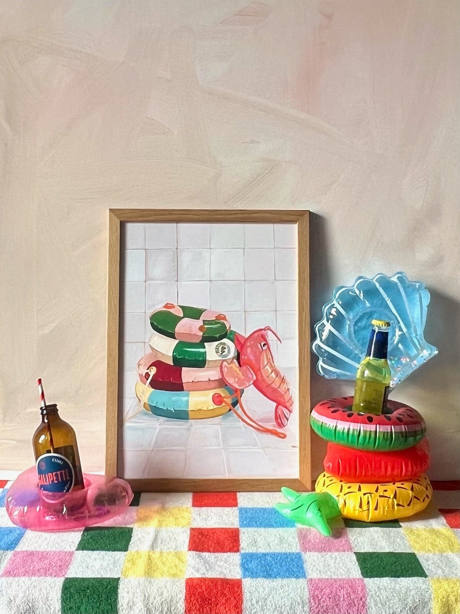 The Platypus Pool Pals Giclée Art Print by Hôtel Dodo, featuring vibrant inflatable pool rings, leans against a wall among drink bottles in inflatable cup holders on a checkered surface.