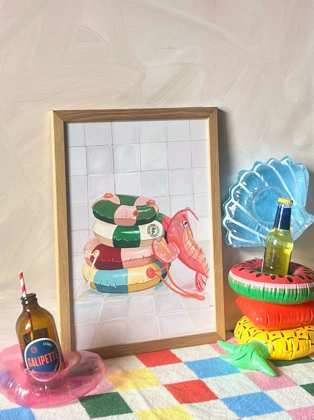 The Platypus Pool Pals Giclée Art Print by Hôtel Dodo leans against a wall, next to bottles in shell, watermelon, and donut pool rings, all arranged on a colorful checkered surface.