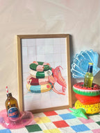 The Platypus Pool Pals Giclée Art Print by Hôtel Dodo leans against a wall, next to bottles in shell, watermelon, and donut pool rings, all arranged on a colorful checkered surface.
