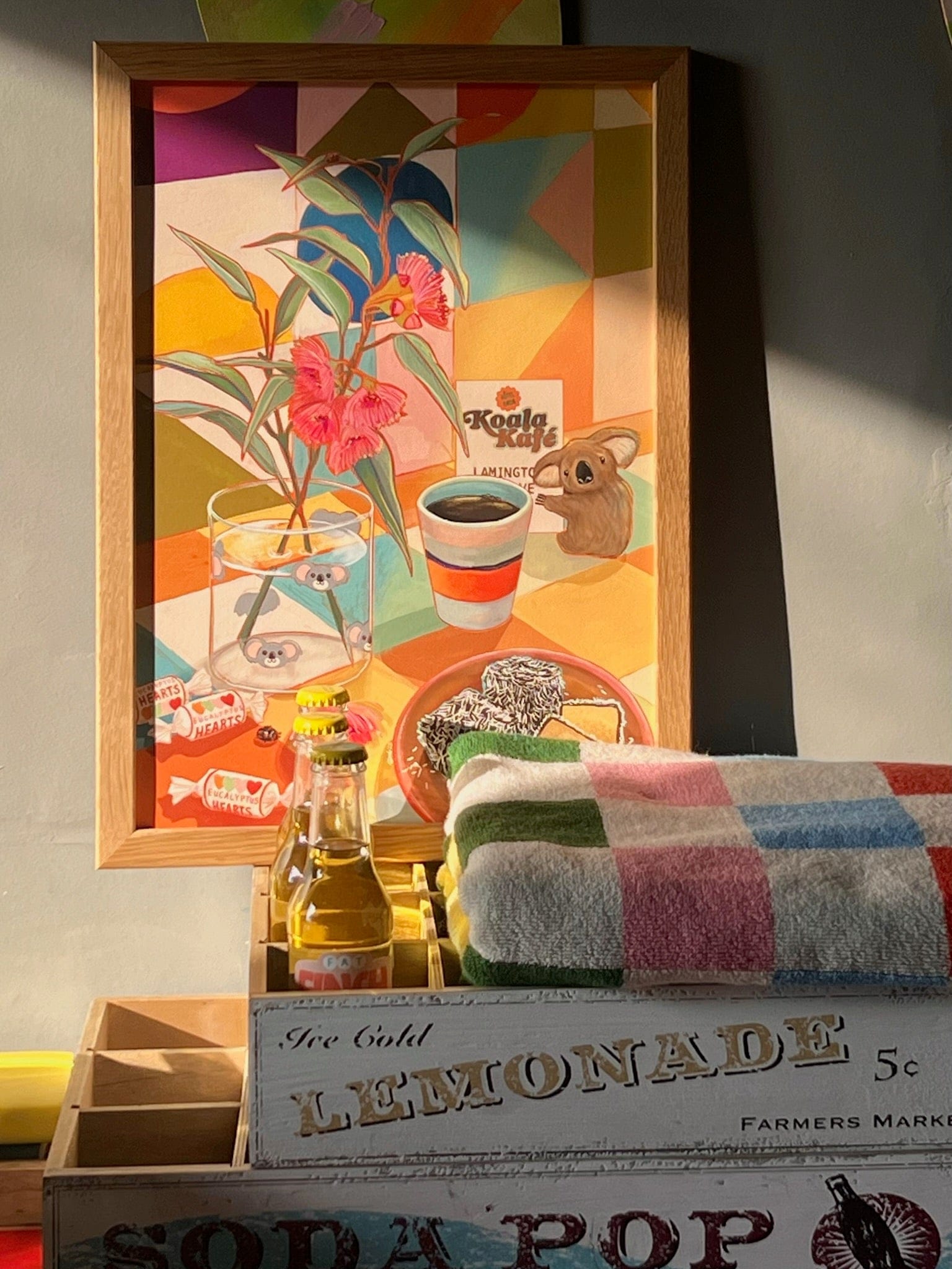 The Koala Kafé Giclée Art Print by Hôtel Dodo hangs above wooden crates labeled "LEMONADE" and "SODA POP," displaying a glass bottle, checkered towel, and retro Australiana decor bathed in sunlight—evoking cozy café vibes.