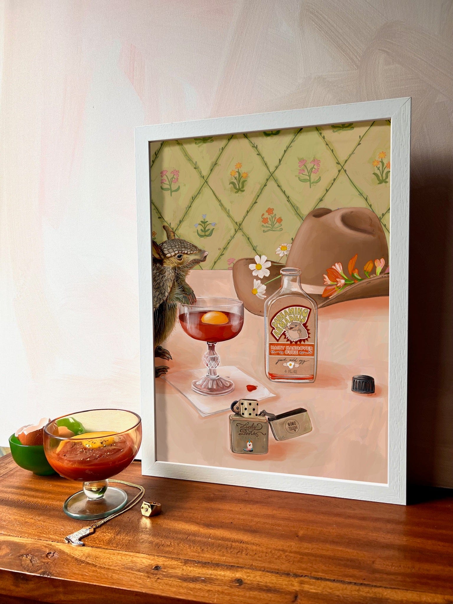 The Prairie Oyster Giclée Art Print by Hôtel Dodo features a vibrant cocktail with an orange slice, whiskey bottle, dice, hat, and small animal—an eye-catching décor piece ideal for any gift shop display.