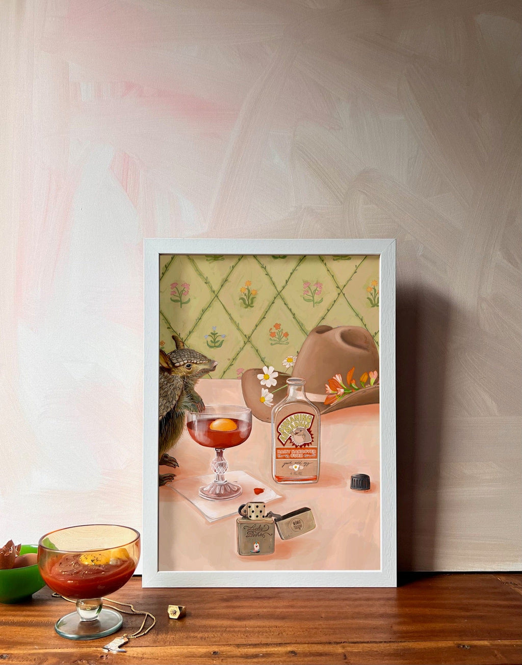The Hôtel Dodo Prairie Oyster Giclée Art Print features a vintage still life with a squirrel, bottle, dice, teapot, and prairie oyster cocktail and is displayed on wood beside a real glass—perfect for an artful gift shop vibe.